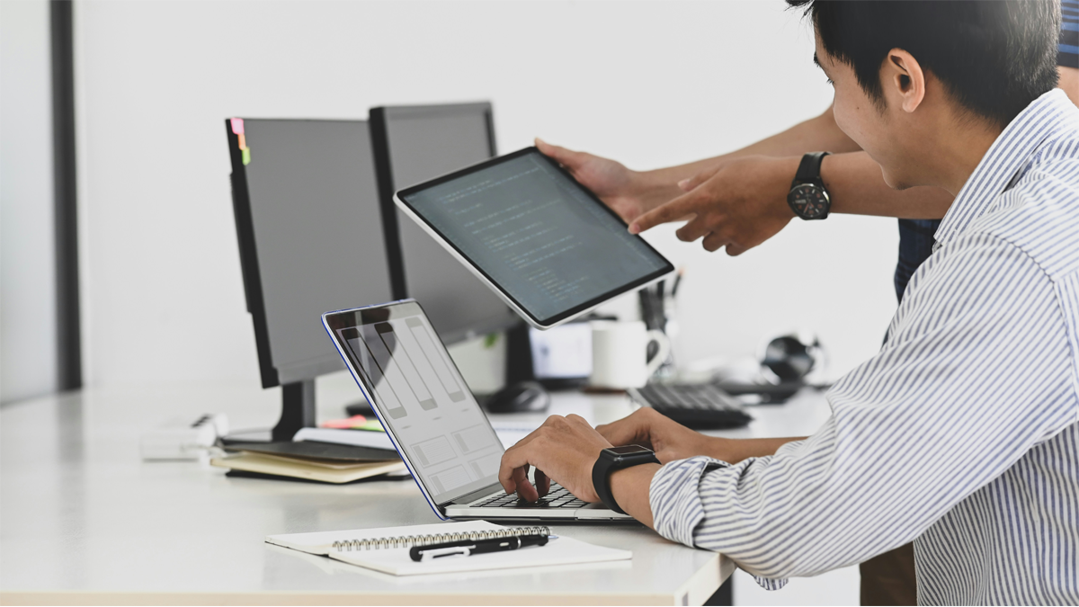 Office workers at desk with multiple tech devices.